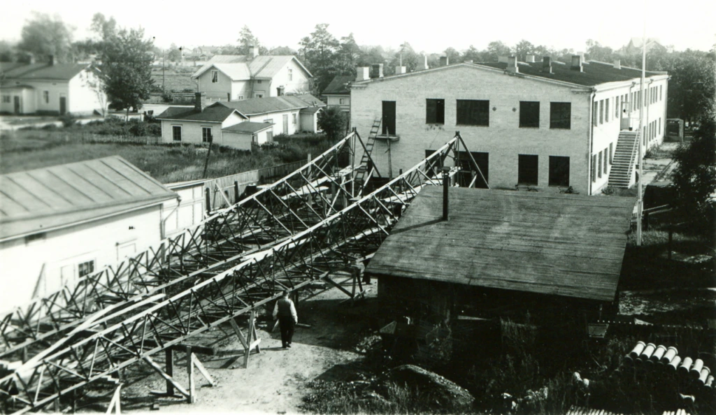 Mannerin konepaja rakentaa radiomaston. Kuvassa näkyy sekä tehdas- että asuinrakennuksia. Manners mekaniska verkstad bygger en radiomast. På bilden både fabriks- och bostadshus. The Manner mechanical workshop buildling a radio mast. There's both factory and dwelling houses in the picture.