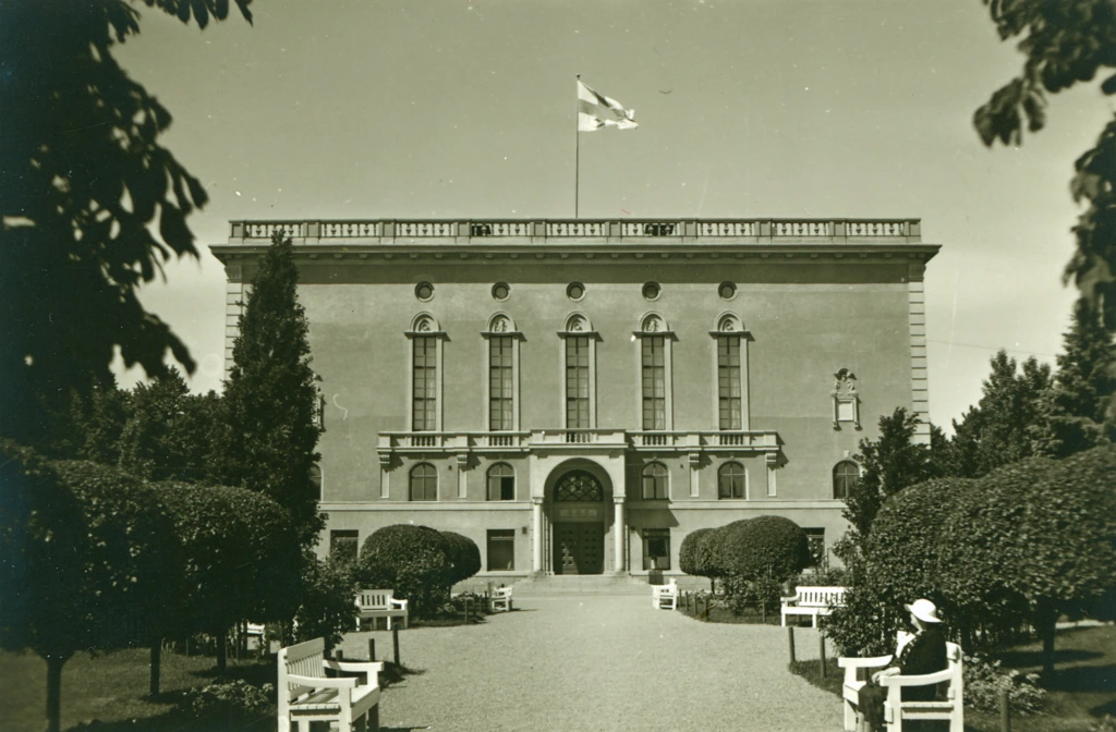 Hangon vanha kaupungintalo ja sen edustalla oleva puisto. Nainen istuu penkillä. Hangös gamla stadshus oh arken framförden. En kvinna sitter på en bänk. The old town hall of Hanko and the park in front of it. A woman sits on a bench.