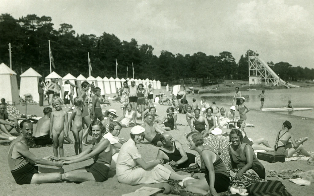 Plagenin ranta 1920-luvulla. Ranta on täynnä ihmisiä ja uimakoppeja. Taustalla korkea liukumäki. Plagenstranden på 1920-talet. Strand är fylld med människor och badhyttar. I bakgrunden en hög rutschkana. The Plagen beach in the 1920s. The beach is filled with people and beach cabins. A tall slide in the background.