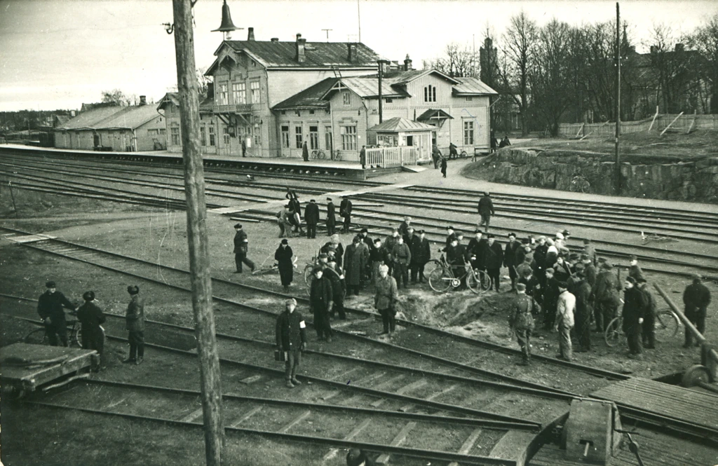 Hankolaiset ovat kokoontuneet katsomaan pommin tekemää kuoppaa ratapihalla vuonna 1939. Taustalla näkyy vanha rautatieasema, vanha vesitorni ja muita rakennuksia. Hangöborna står samlade kring en grop som en bomb har lämnat på bangården 1939. I bakgrunden syns den gamla järnvägsstationen, det gamla vattentornet och andra byggnader. Residents of Hanko have gathered around a pit left by a bomb in the railway yard in 939. In the background you can see the old railway statoin, the old water tower and other buildlings.